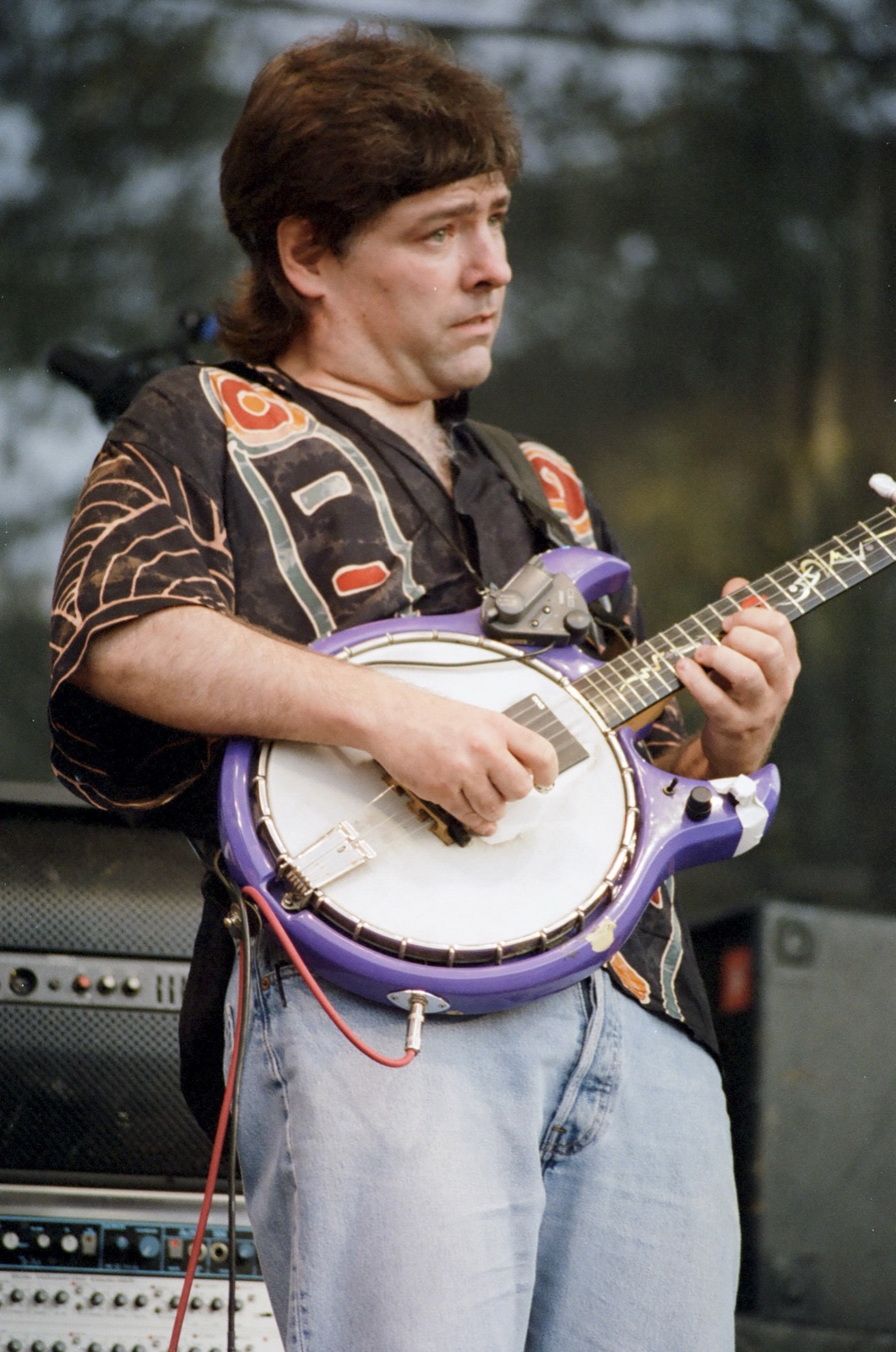 A banjo player in a patterned shirt plays a purple electric banjo on stage.